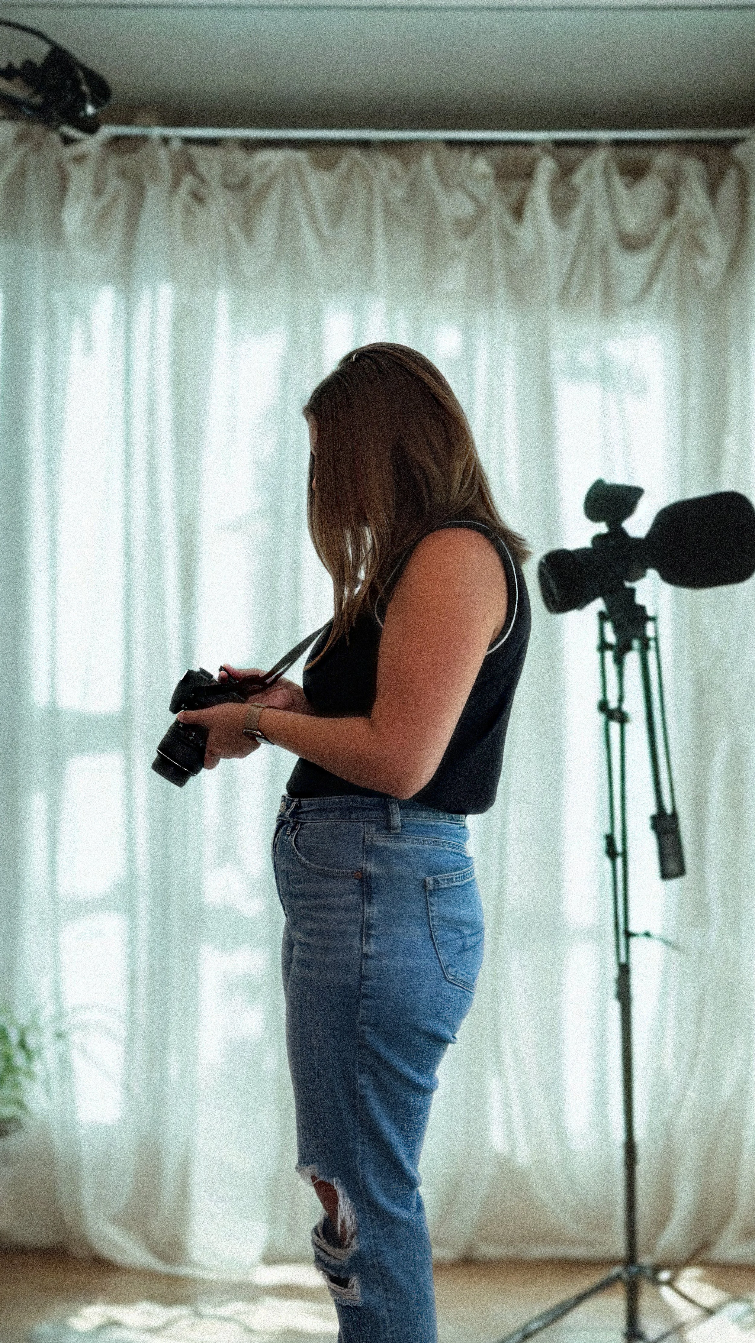 A woman with brown hair, wearing a sleeveless black top and ripped blue jeans, holds a camera while standing indoors in front of a window with white curtains. A professional camera on a tripod is nearby.