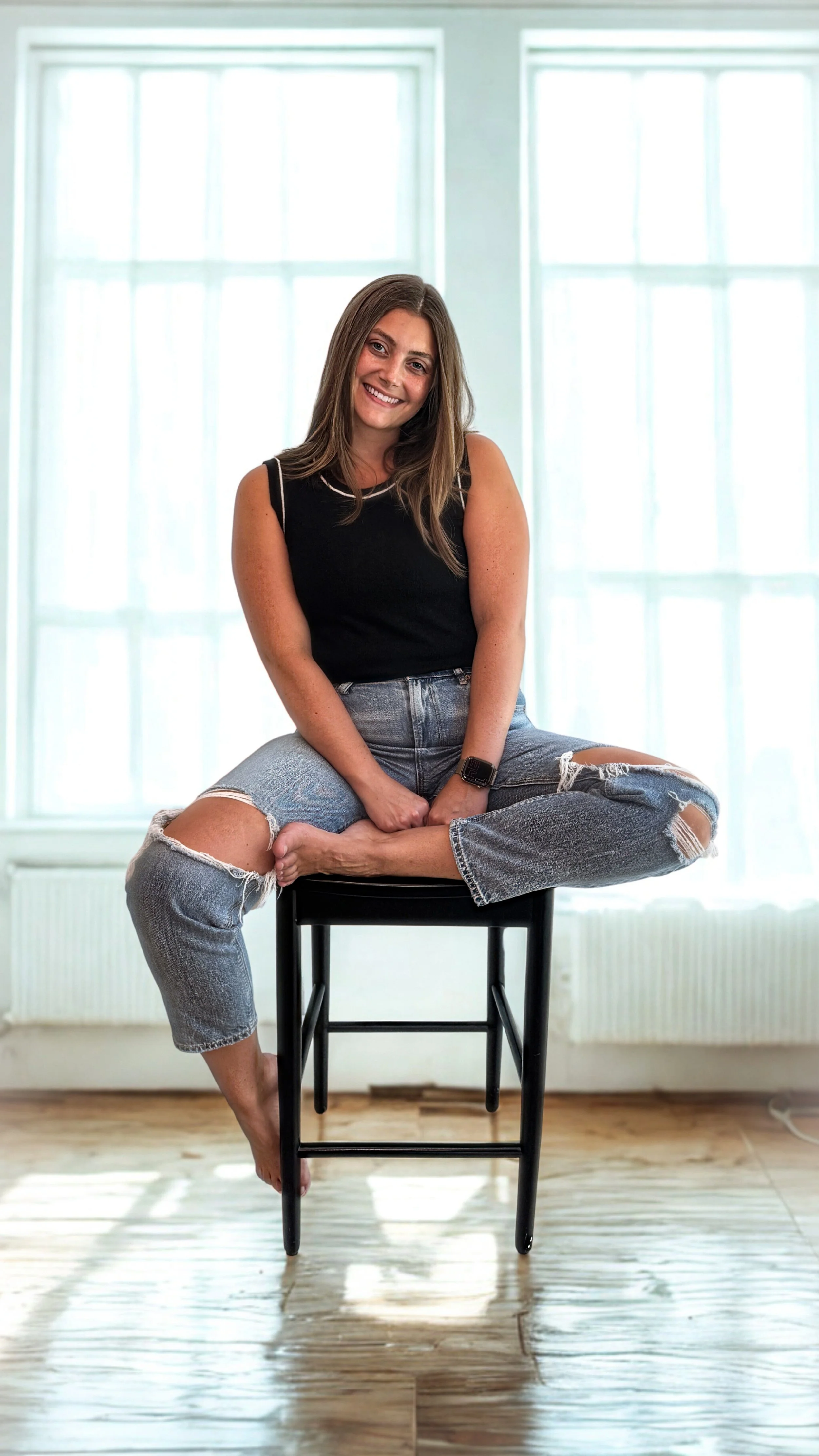 A woman with long brown hair sitting cross-legged on a black stool in front of large windows, smiling at the camera, wearing a black top, ripped jeans, and a smartwatch in a bright room.