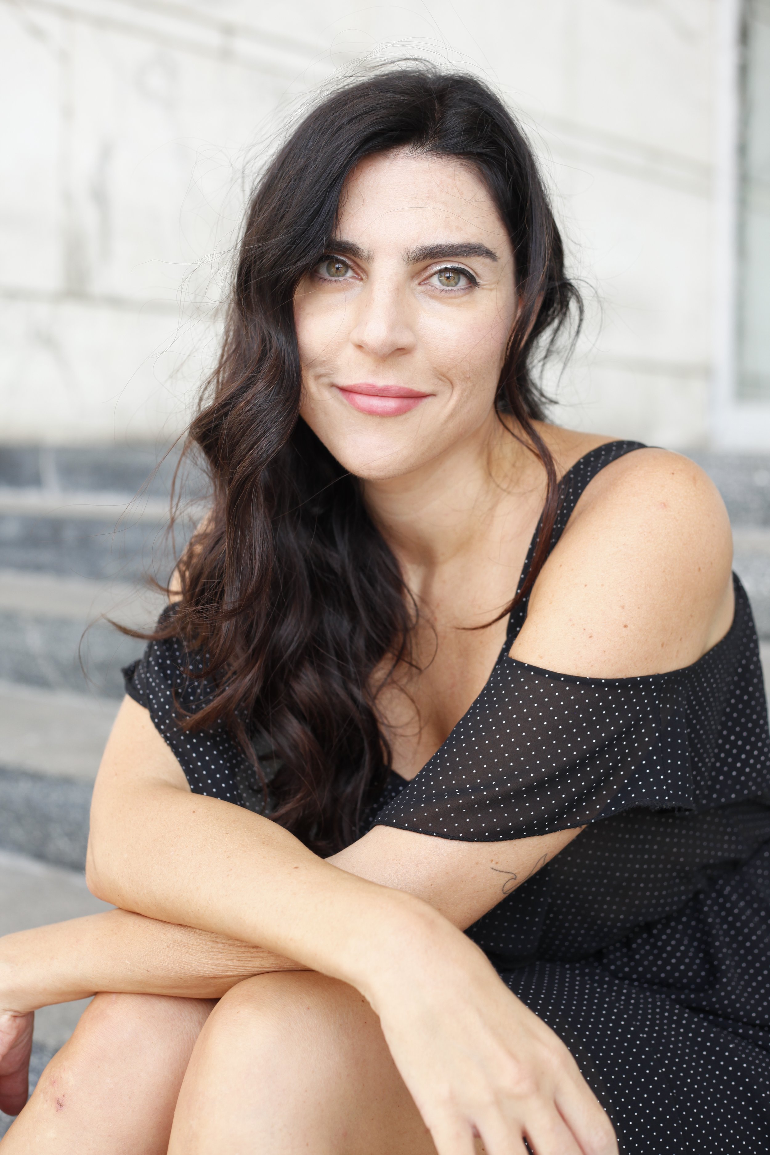Donna con capelli castani ricci e occhi chiari, vestita con un abito nero a pois bianchi, seduta sulle scale in pietra, con un sorriso.