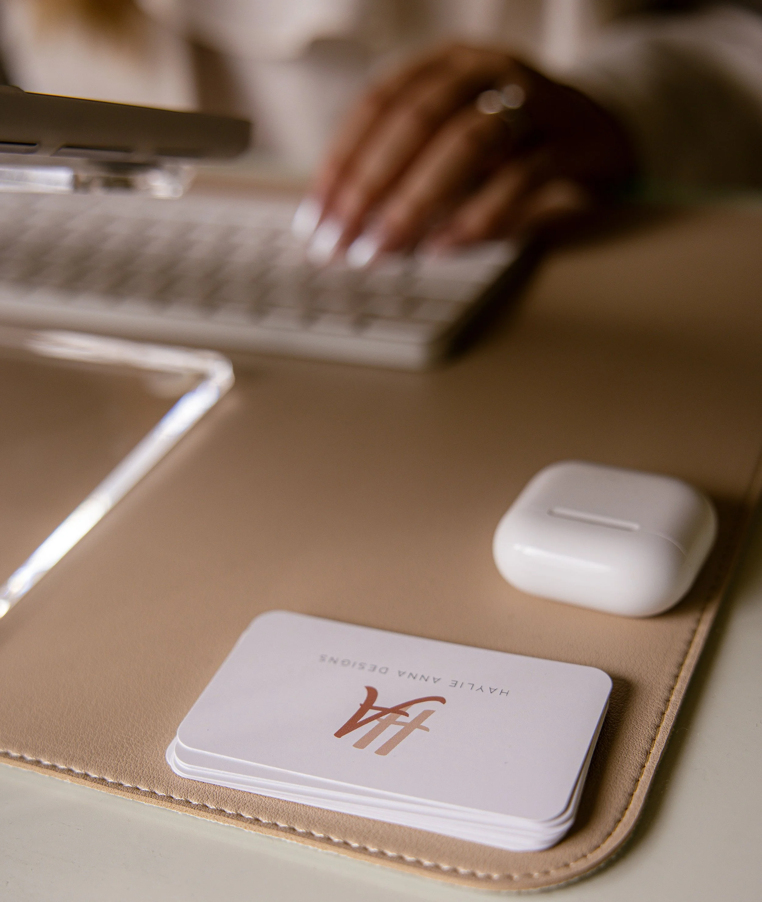 Business cards, AirPods, and a laptop on a beige desk mat, with a blurred hand working on a laptop in the background.