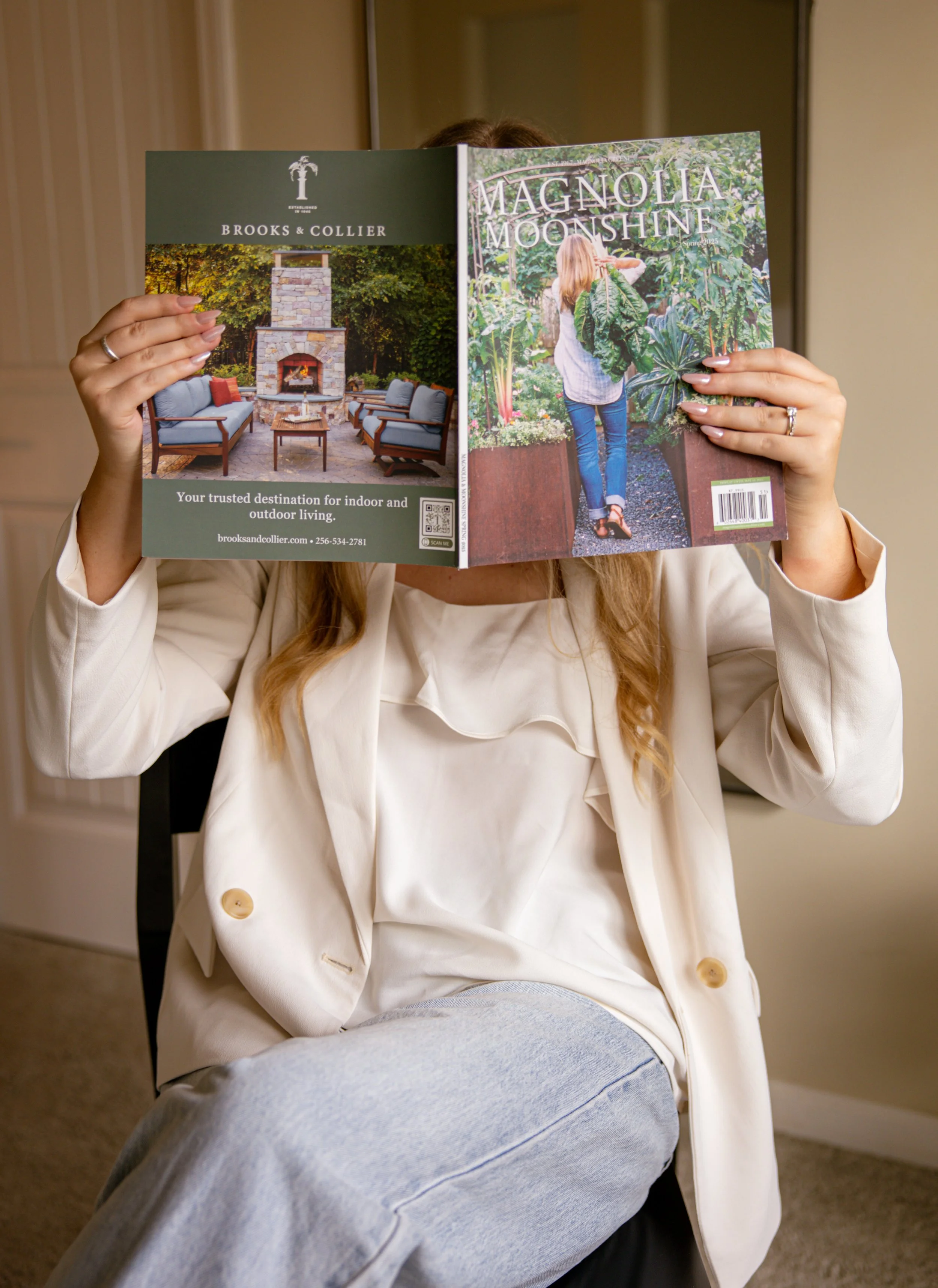 A woman in a white blazer and light gray pants is sitting while reading a magazine titled "Magnolia Moonshine." The magazine features an outdoor garden scene with a woman in a white blouse and jeans on the cover.