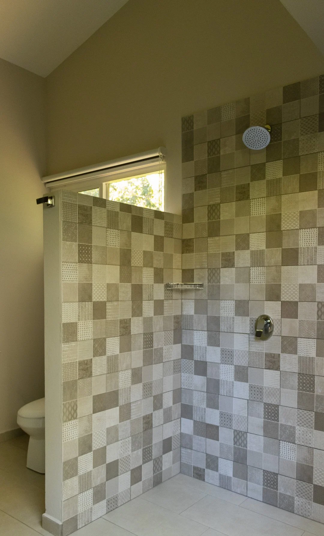 A modern bathroom with a tiled walk-in shower featuring beige and brown patterned tiles, a round showerhead, a small window above the shower, and a partial view of a toilet in the corner.