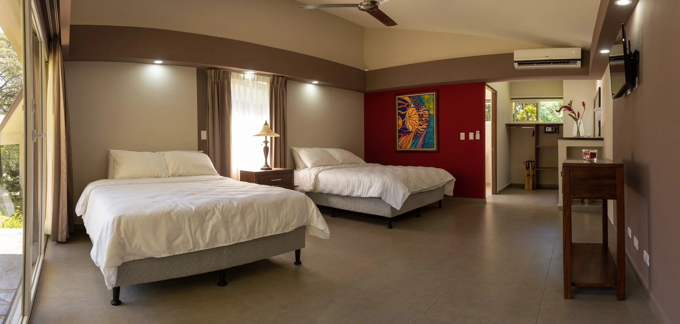 A hotel room with two beds, nightstand, lamp, sliding glass door, window with curtains, wall art, small open closet, and mounted TV, with beige and red walls
