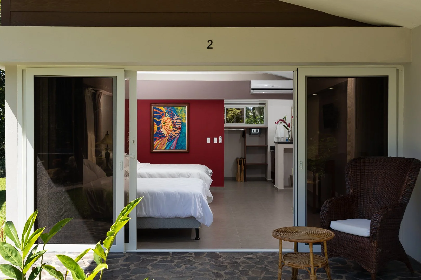 View from outside a sliding glass door into a modern bedroom with a red accent wall, colorful abstract painting, white beds, and a small kitchenette area with a coffee maker and flowers.