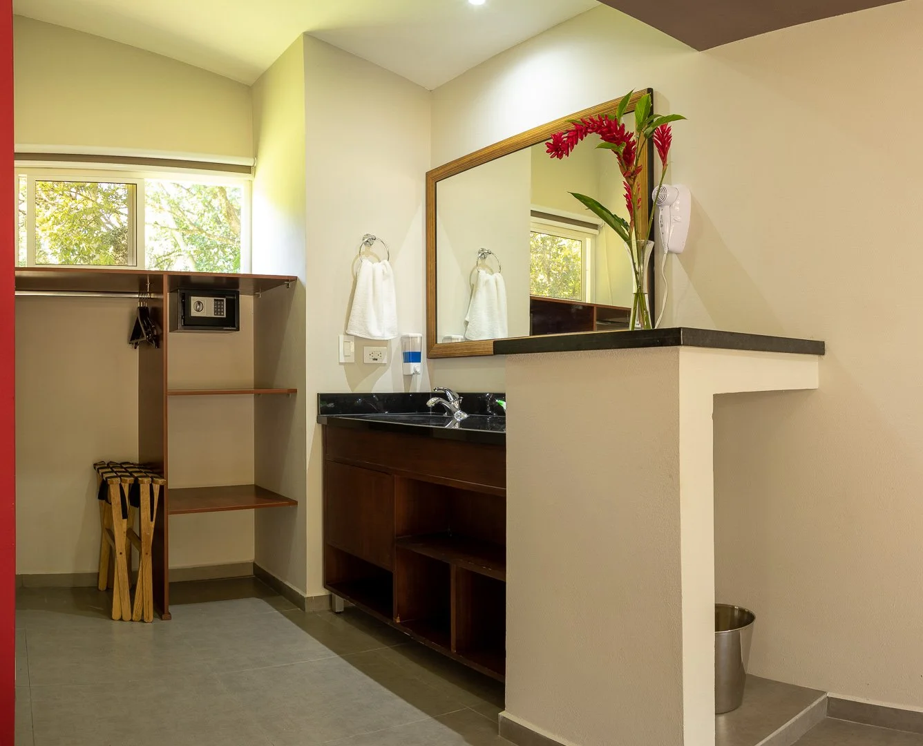Hotel room bathroom with a mirror, flowers, towels, a sink, and an open closet with a safe.