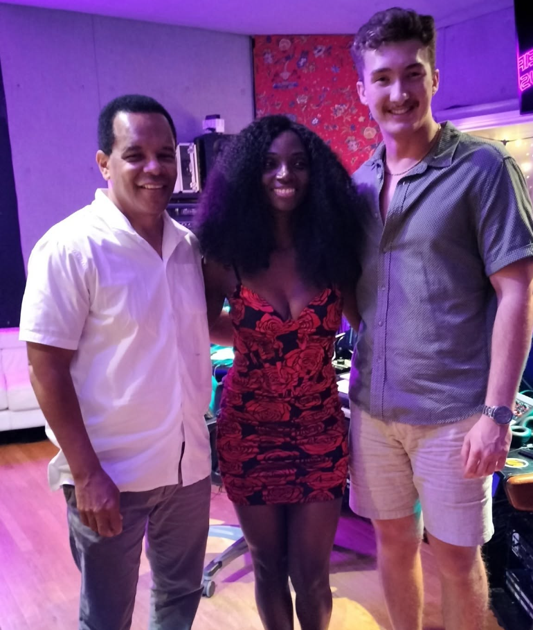 Three people, two men and one woman, standing together in a colorful indoor setting with purple lighting, smiling at the camera.