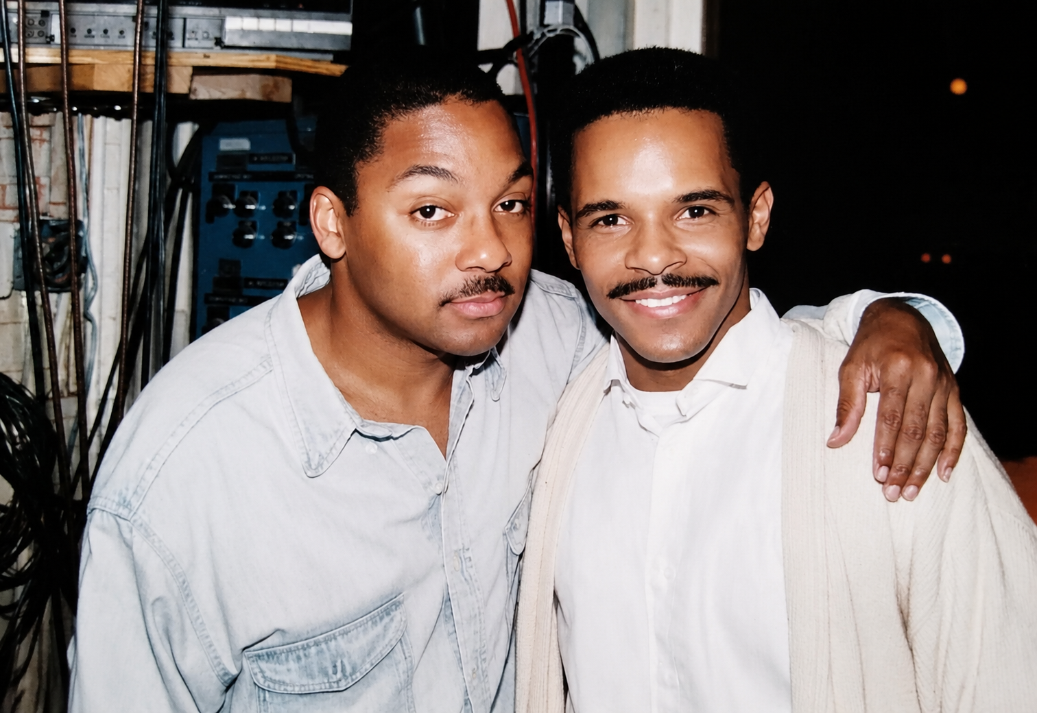 Two men with dark hair and mustaches smiling, one with a white shirt and the other with a cream-colored cardigan, standing close together with the man in the cardigan having his arm around the other's shoulder.
