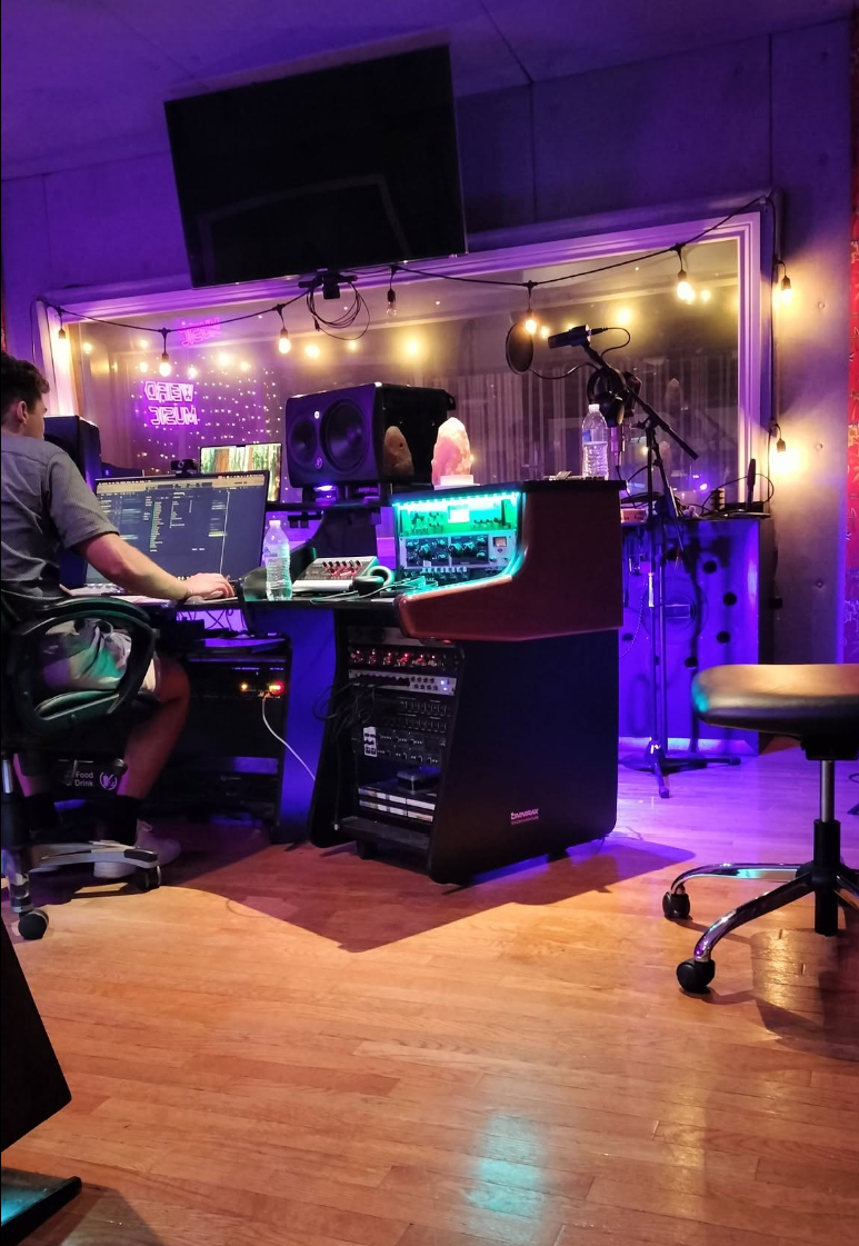 Music recording studio with electronic equipment, monitors, and microphones, illuminated by colorful purple and yellow lighting.