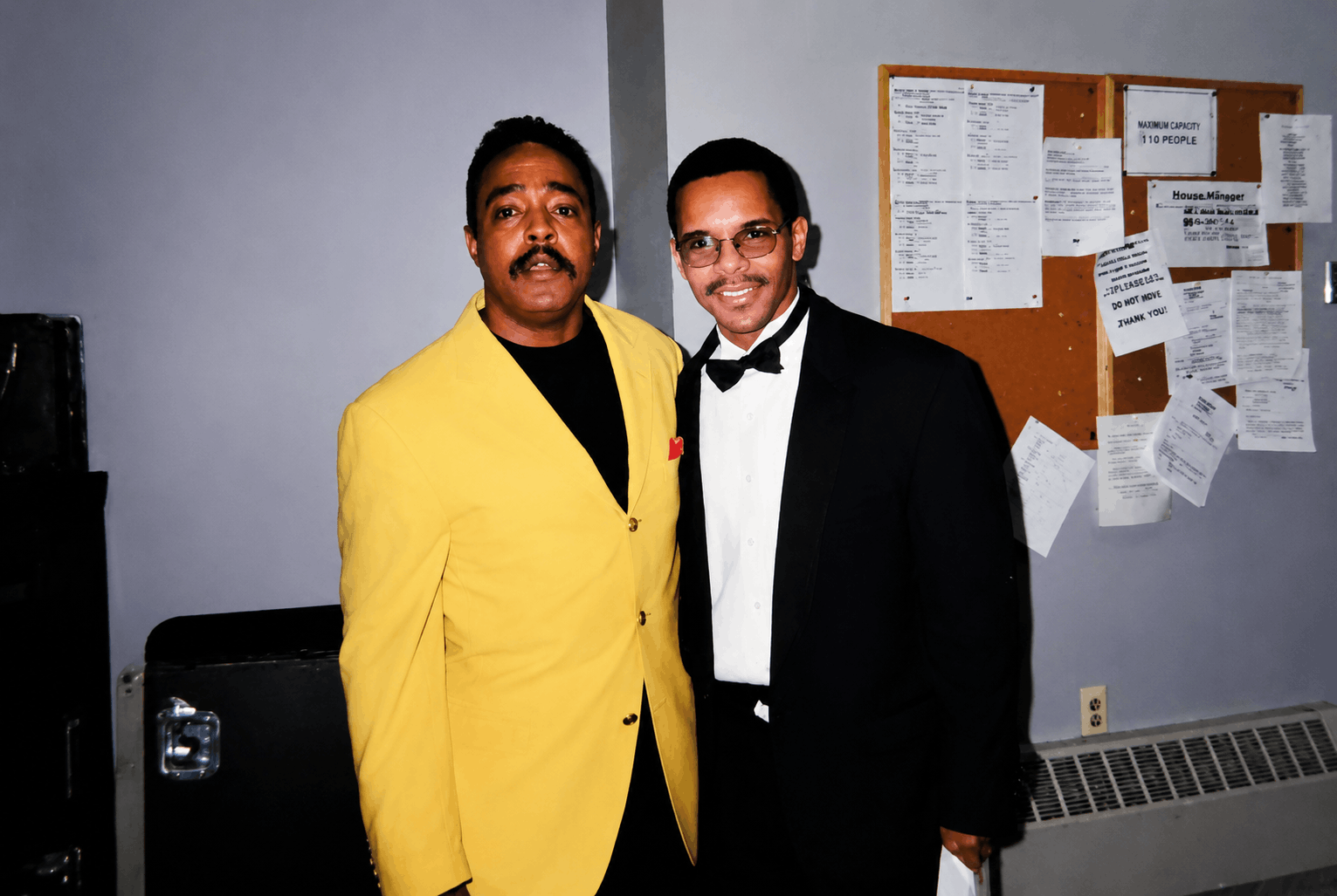 Two men standing close together, one wearing a bright yellow suit and the other in a black tuxedo with a bow tie, posing for the photo in an indoor setting with a bulletin board behind them. Peebo Bryson. Music star. Grammy. Grammys. Music Manager.