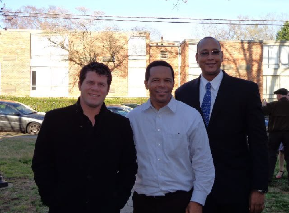 Three men dressed in formal attire, standing outdoors with a brick building, trees, cars, and other people in the background. Music awards. Award show.