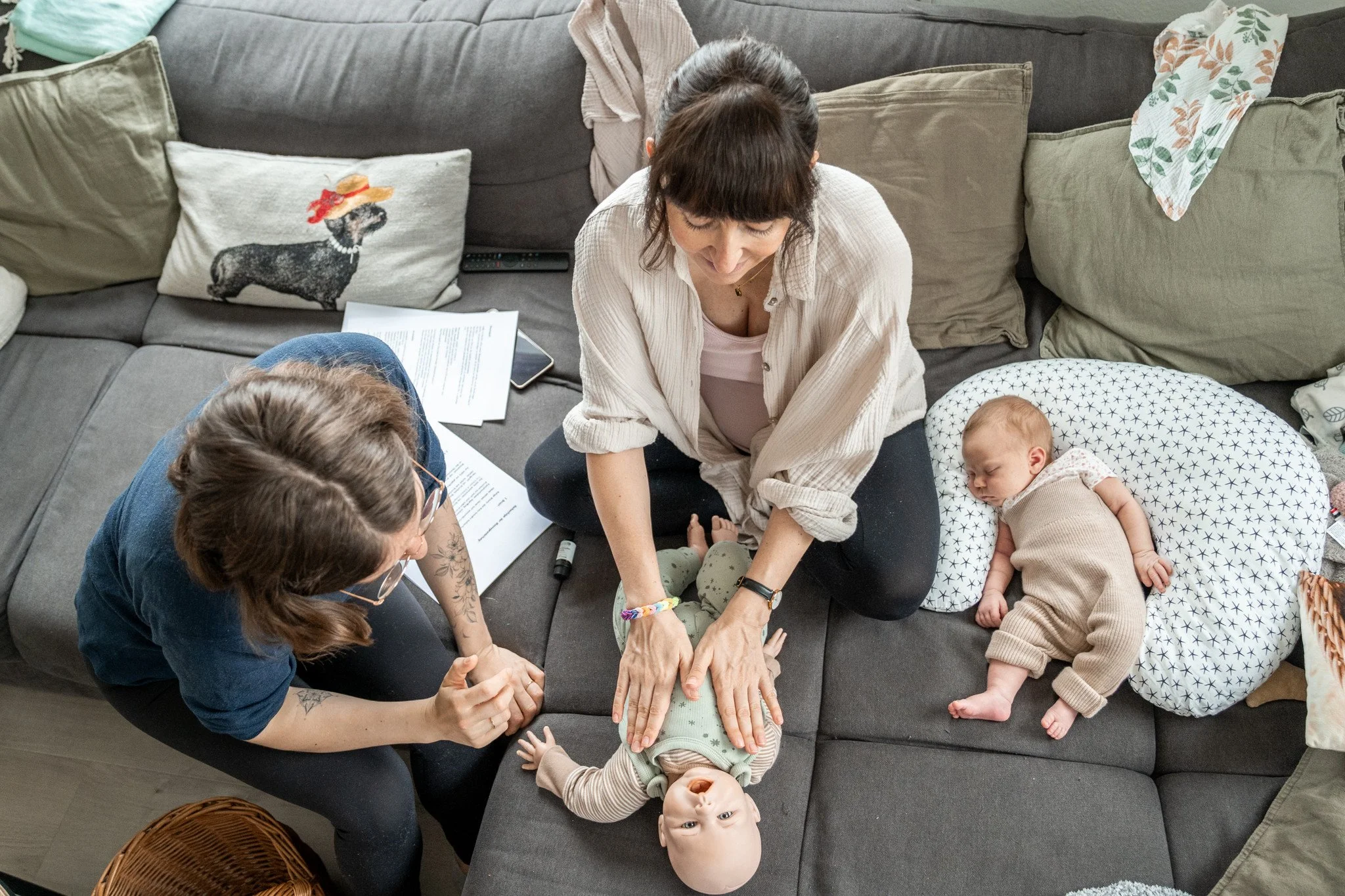 Auf diesem Bild zeige ich einer Mama, wie sie ihr Baby massieren kann. Wir sitzen auf einem Sofa und üben dabei an einer Puppe. Ihr Baby schläft auf einem Kissen neben ihr.