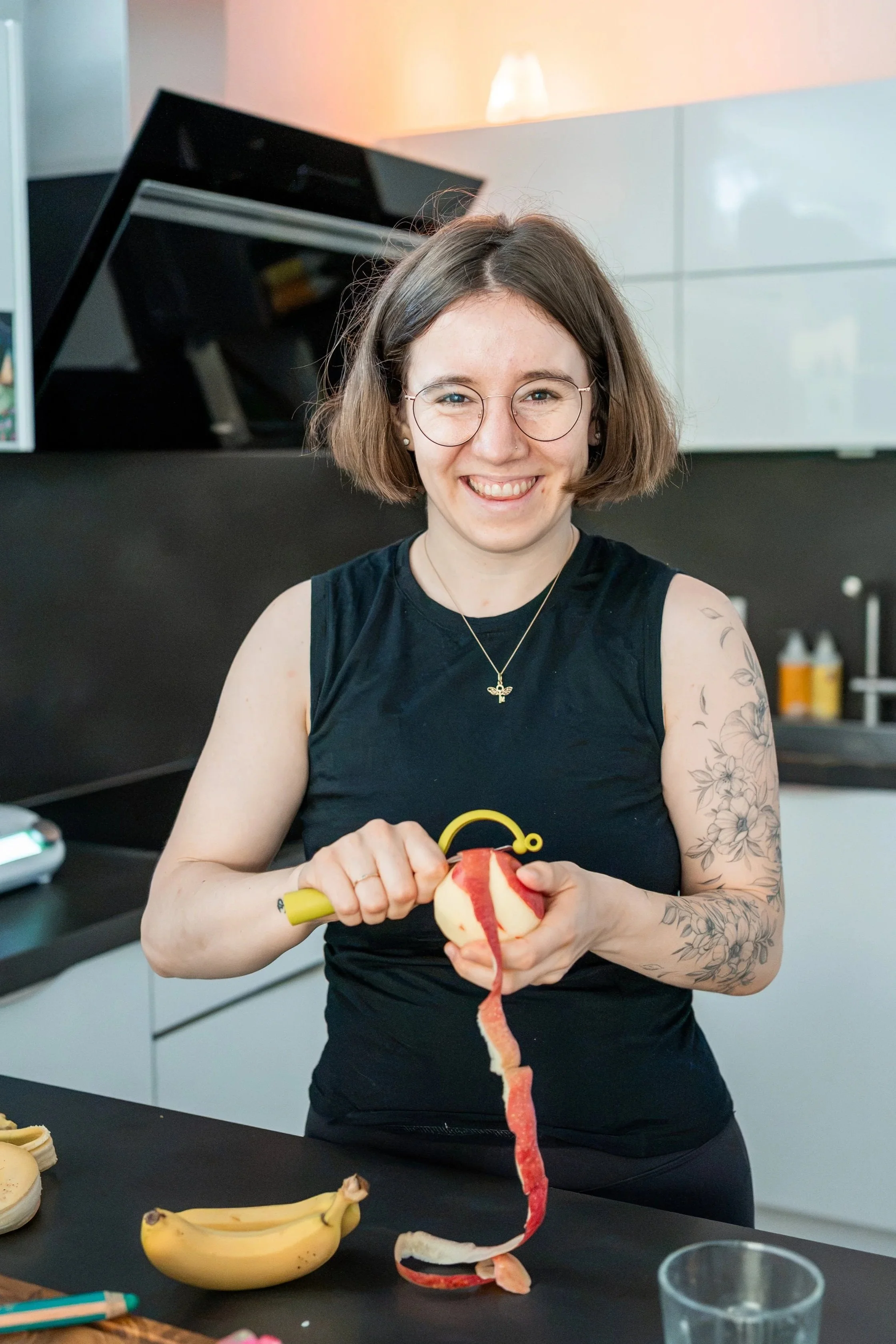 Auf diesem Bild schäle ich gerade einen Apfel. Ich habe kinnlange, braune Haare, trage eine goldene Brille und habe ein Fineline-Tattoo mit Blumen auf dem Arm. Ich lächle in die Kamera, im Hintergrund ist eine weiße Küche mit schwarzer Arbeitsplatte.