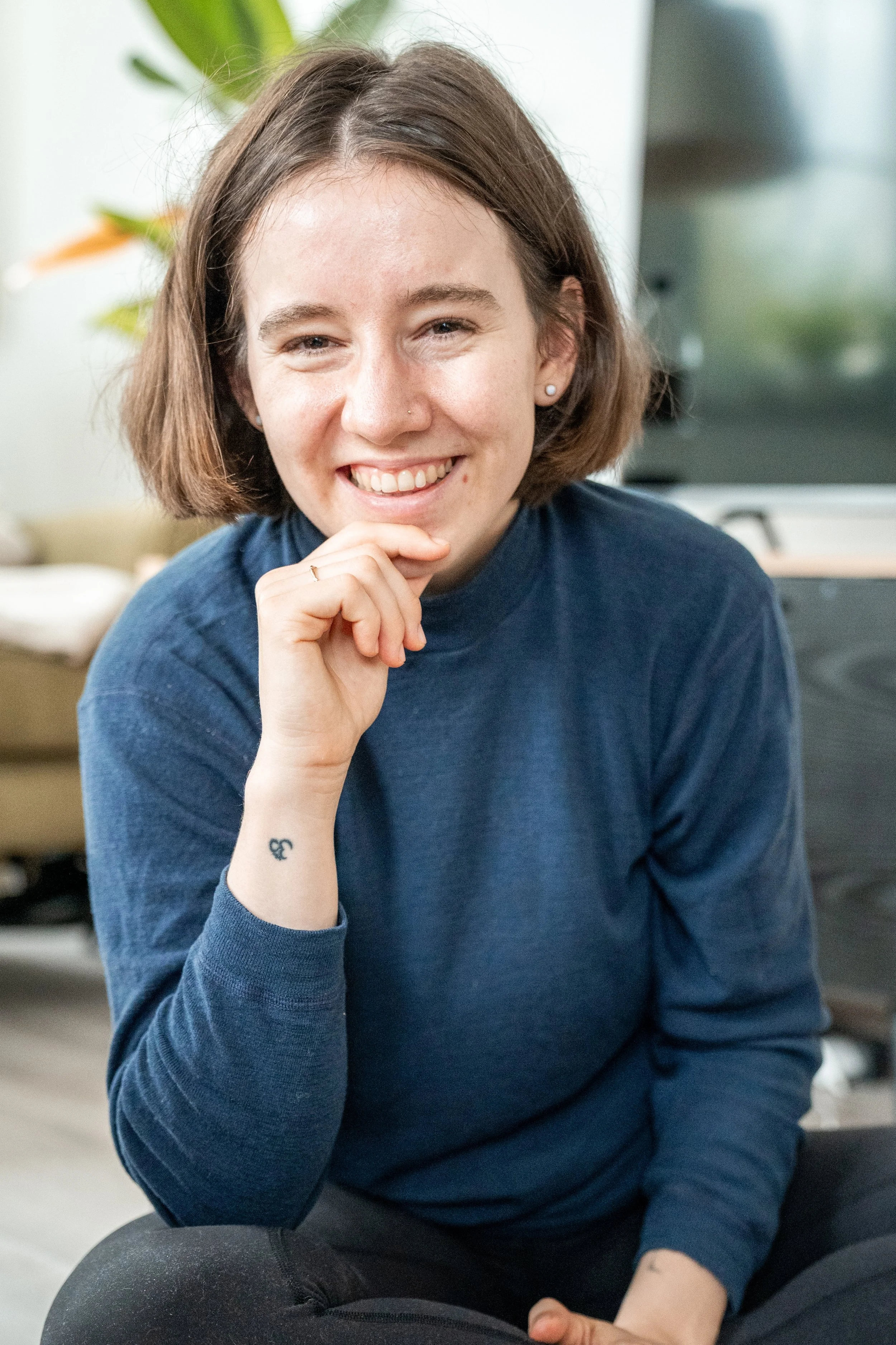 Auf diesem Bild bin du mich, Sophia Morawitz. Ich bin um die 30, habe ungefähr kinnlange braune Haare, trage einen blauen Pullover und lächle in die Kamera. Meine Hand liegt an meinem Kinn und im Hintergrund ist ein Fernseher und eine Pflanze.