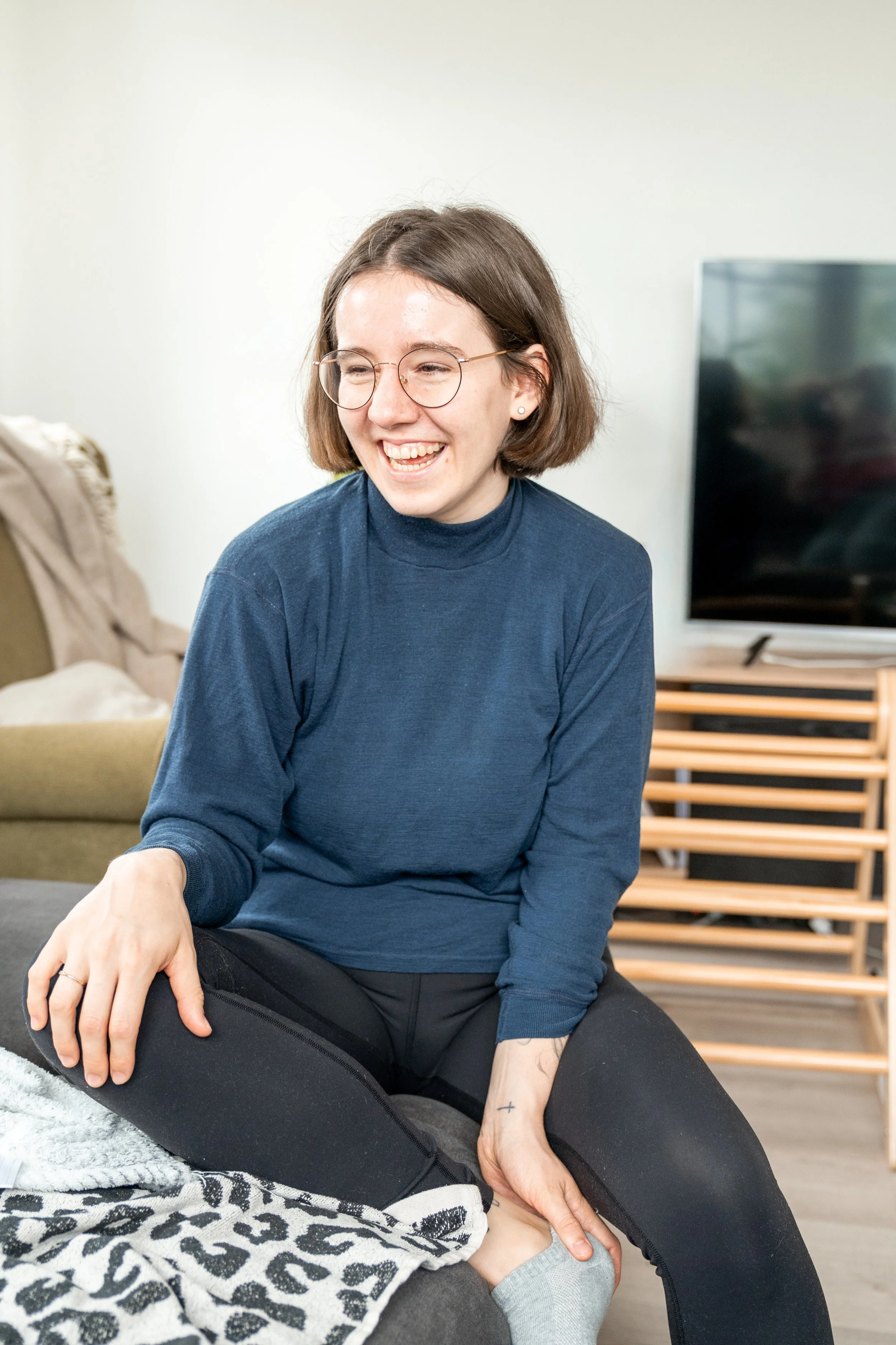 Auf diesem Bild bin du mich, Sophia Morawitz. Ich sitze auf einem Sofa und bin in ein Gespräch vertieft. Ich habe ungefähr kinnlange braune Haare, trage einen blauen Pullover. Im Hintergrund sind ein Fernseher und eine Pflanze auf einem Sideboard.
