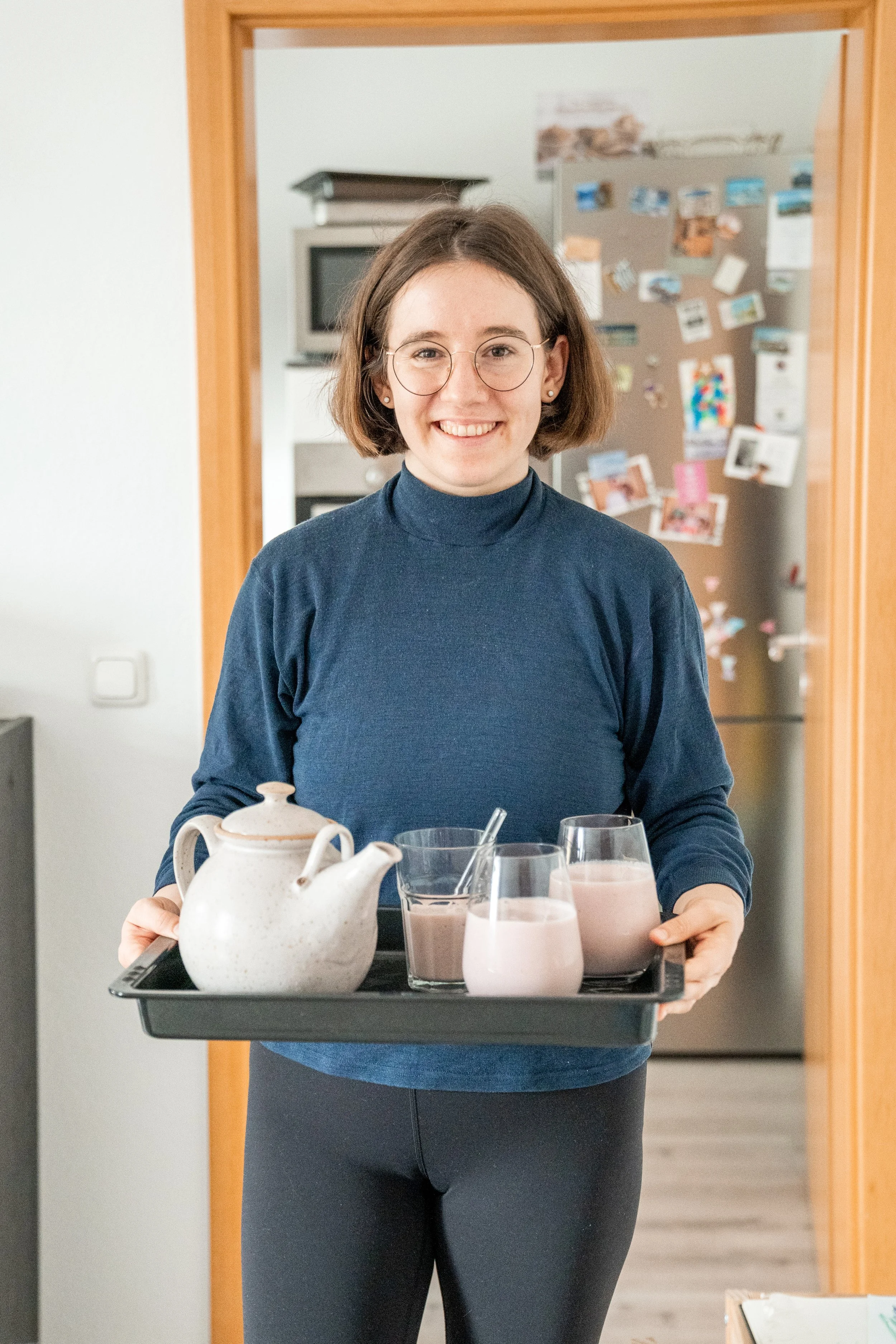 Auf diesem Bild siehst du mich dabei, wie ich gerade eine Erdbeermilch in einer Teekanne mit einem Backblech als Tablett und drei Gläsern darauf ins Wohnzimmer trage. Ich habe einen blauen Pulli und eine schwarze Hose an und lächle in die Kamera.