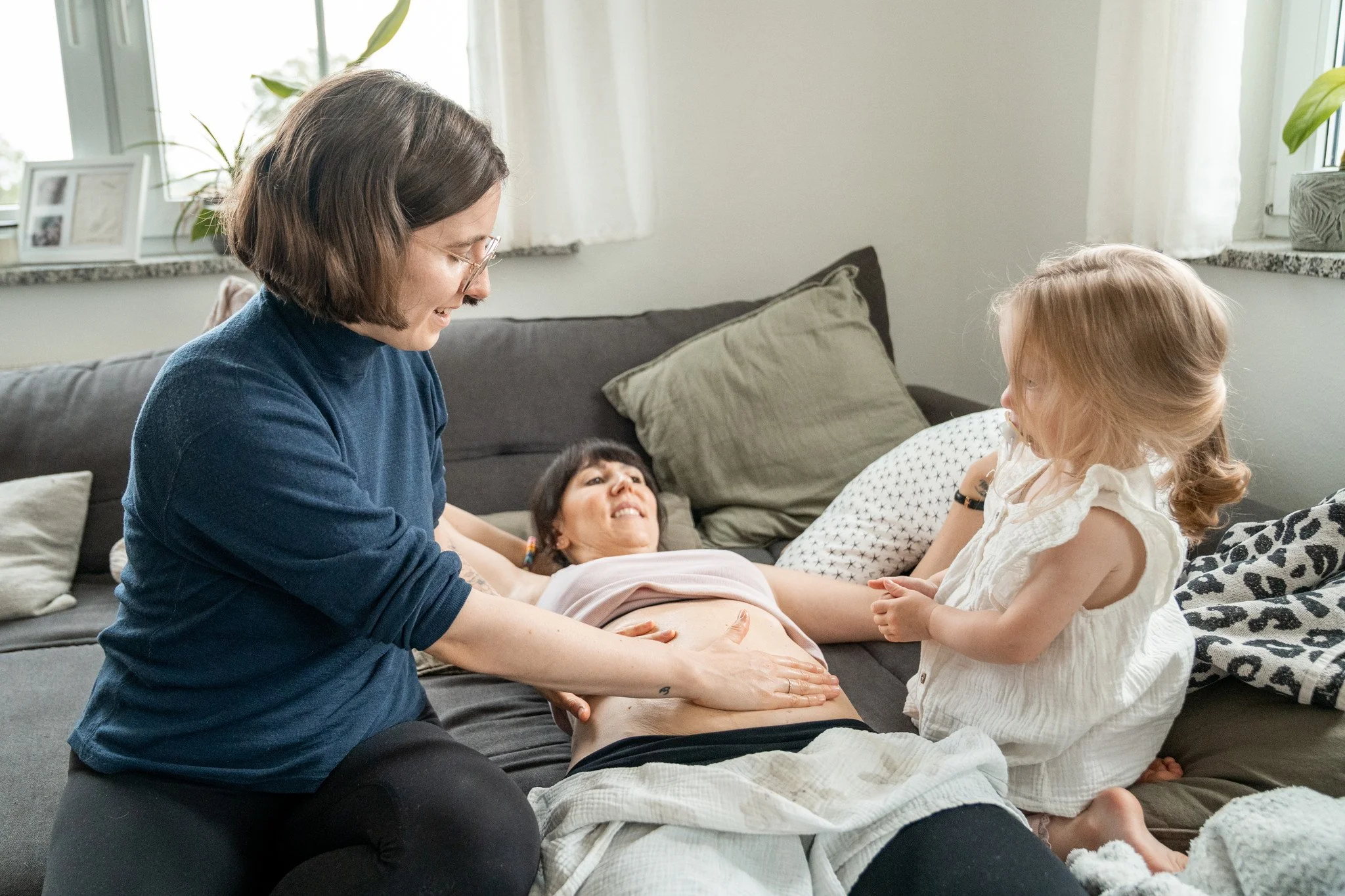Auf diesem Bild massiere ich gerade den Bauch von der Mama, die ich begleite. Sie liegt auf dem Sofa, ich sitze neben ihr auf ihrer rechten Seite, auf ihrer linken Seite sitzt ihre Tochter und ich zeige ihr, wie man Mamas Bauch massieren kann.