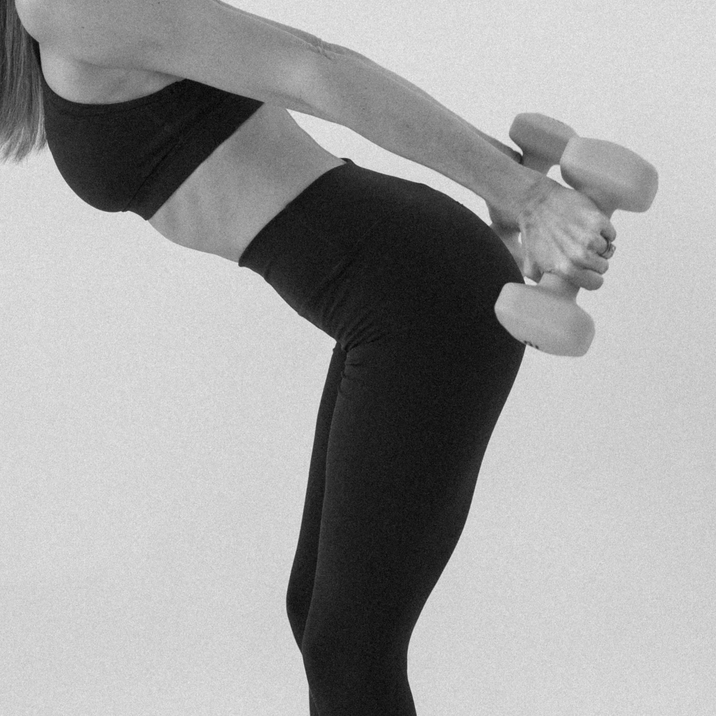 Person holding dumbbells doing a bent-over exercise, wearing black leggings and a sports bra, in black and white.