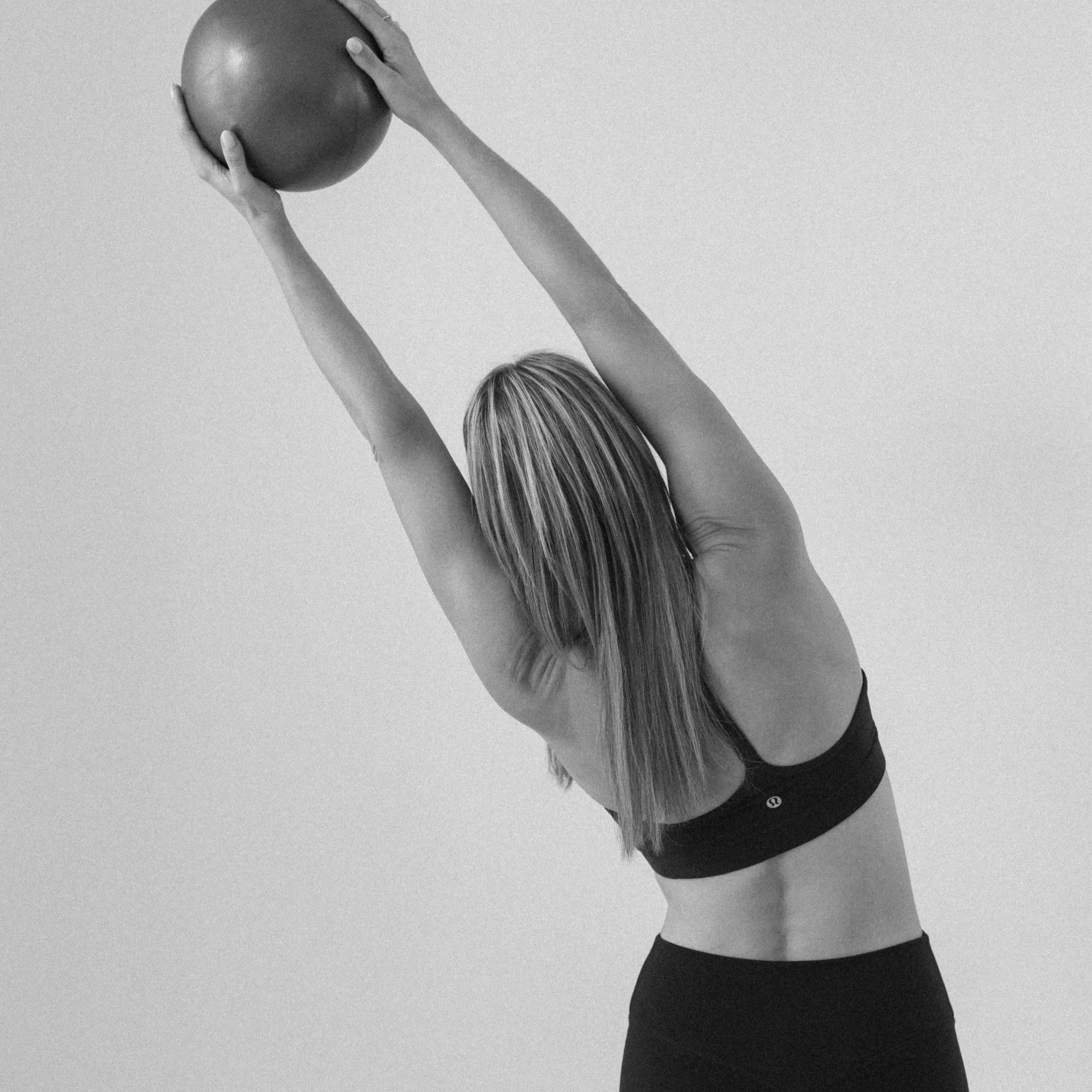 Woman with long hair in sports attire stretching and holding a fitness ball above her head.