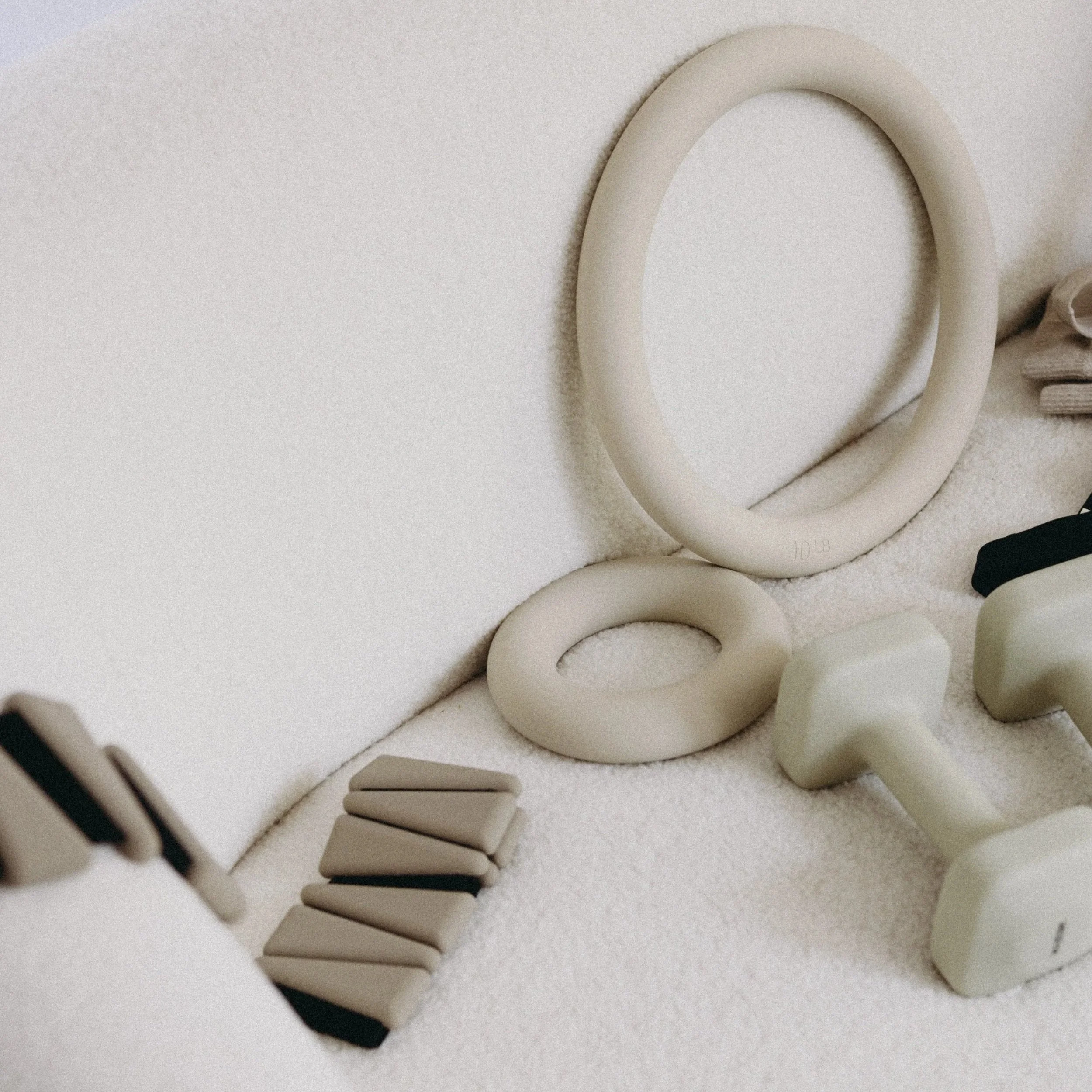 Exercise equipment including a beige fitness ring, a smaller ring, a dumbbell, weight plates, and resistance bands arranged on a soft, textured surface.