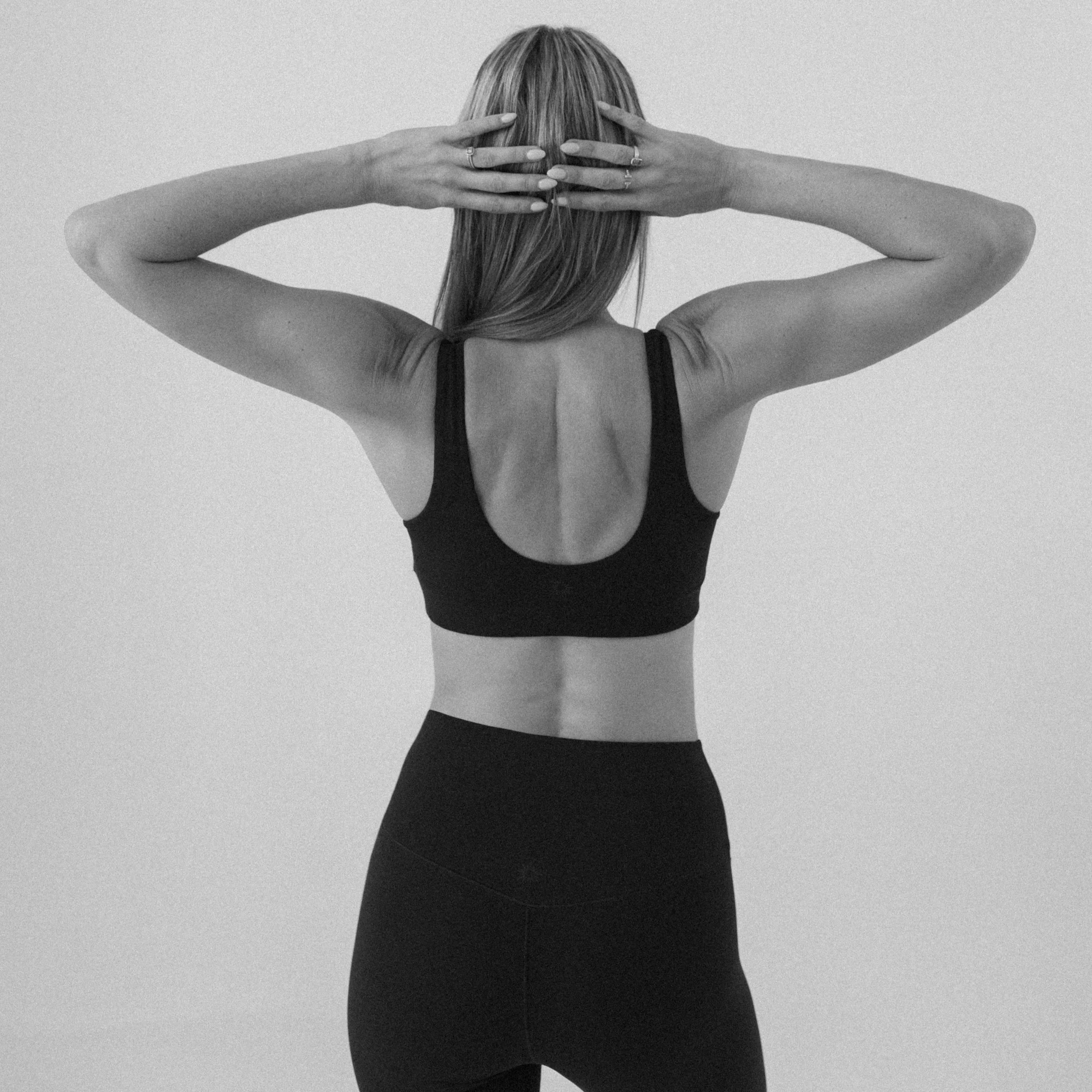A woman in sportswear stretching her arms behind her head against a plain background.