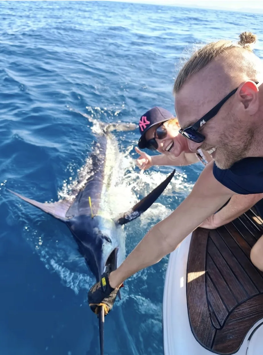 Two people on a boat capturing a selfie with a large marlin fish. The boat is in the ocean, and the sky is clear.