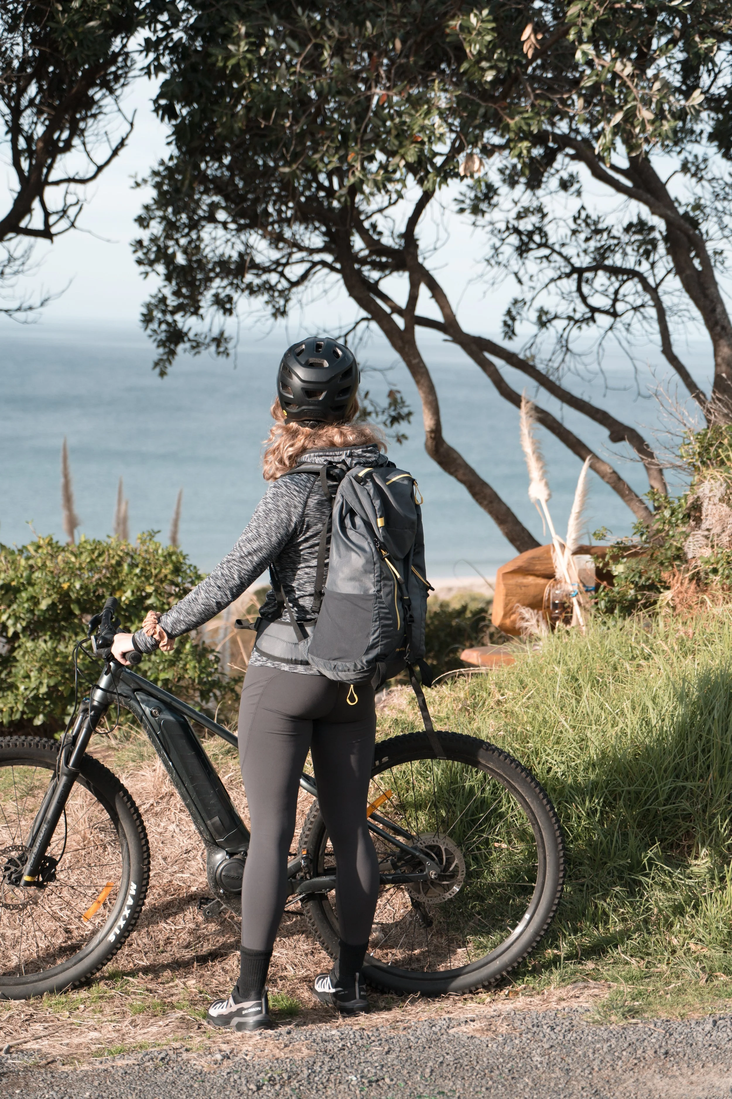 A woman in athletic gear, wearing a helmet and backpack, is standing next to her mountain bike on a grassy trail with a coastal view behind her.