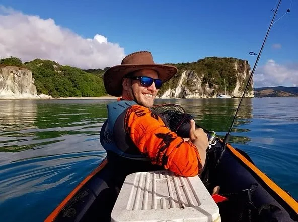 A man in a kayak holding a fishing rod, wearing a wide-brimmed hat, sunglasses, and a life jacket, with a backdrop of cliffs and blue sky.