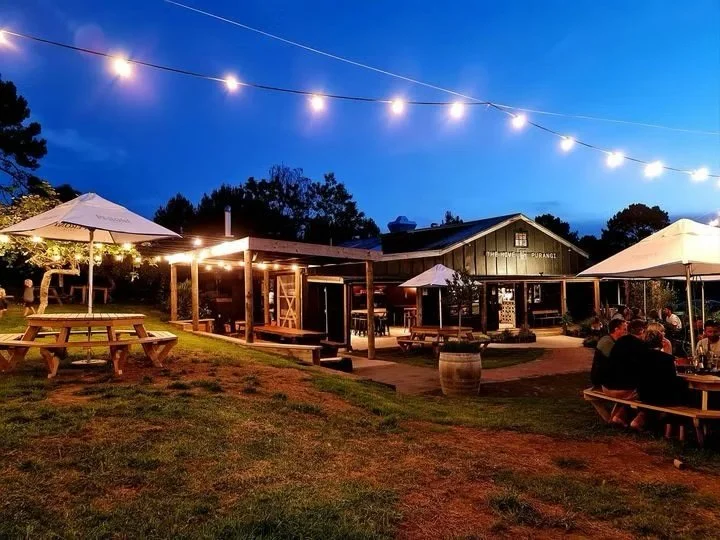 Outdoor dining area at a restaurant during evening with string lights overhead, picnic tables, umbrellas, and people seated at tables.