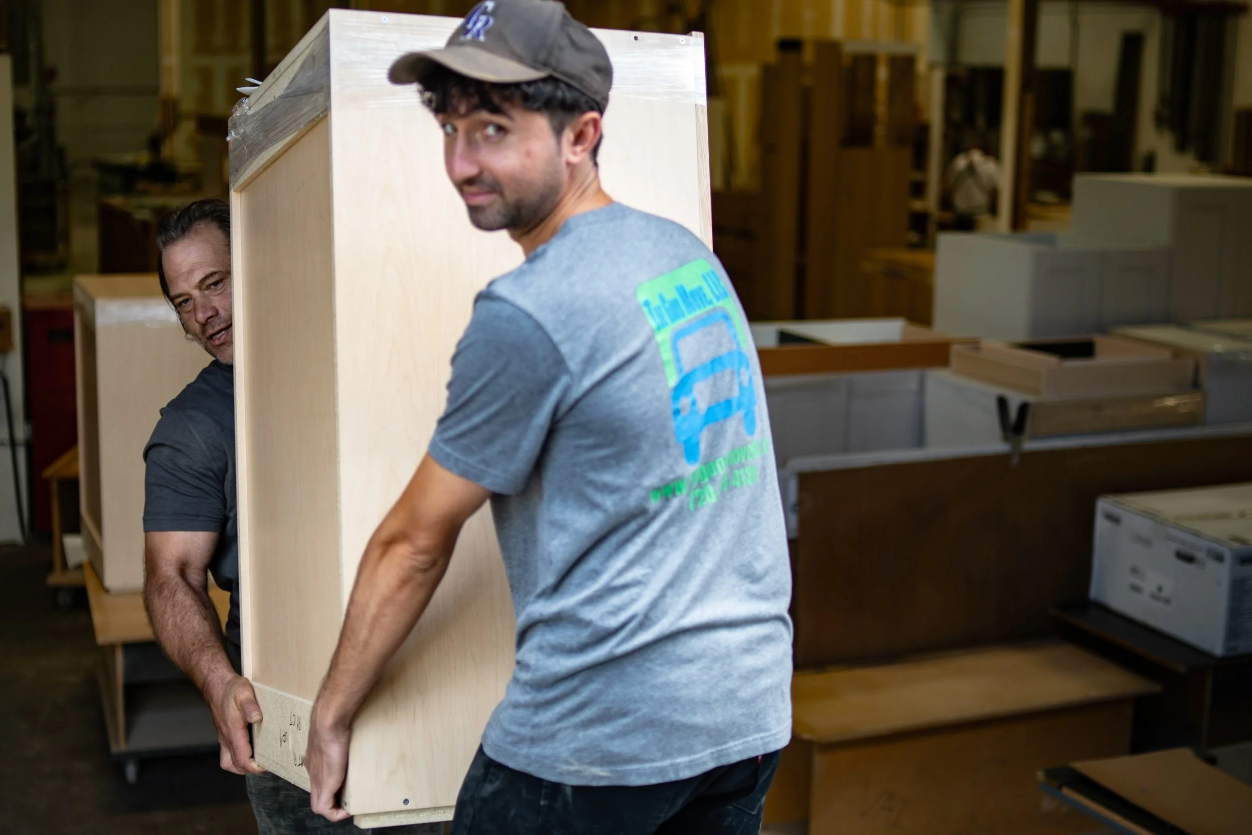Two professional movers in The Good Move shirts lift a large wooden cabinet in a busy warehouse surrounded by moving tools and stacked furniture.