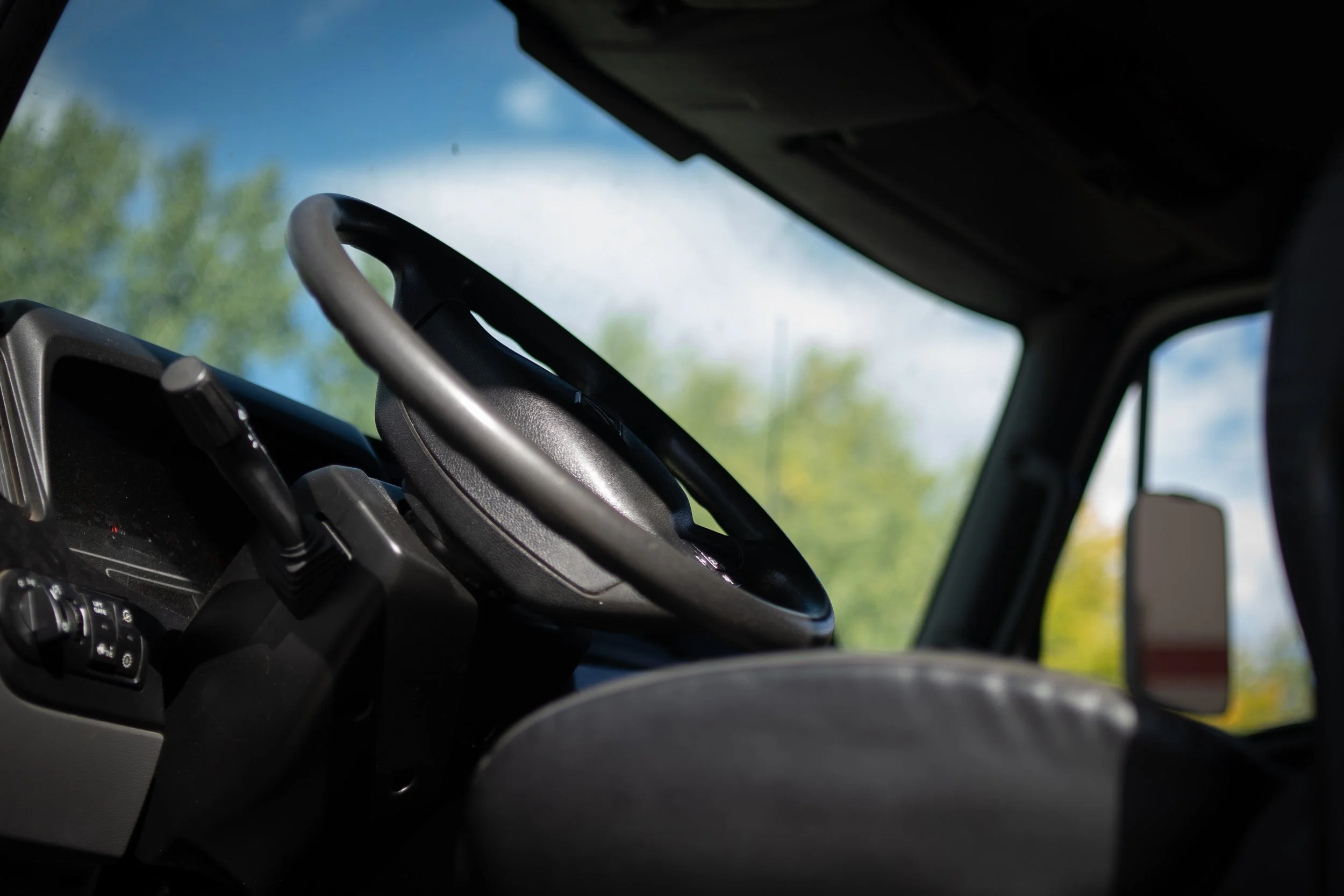 Interior of a vehicle showing the steering wheel, dashboard, and side mirror, with trees and sky visible through the windows.