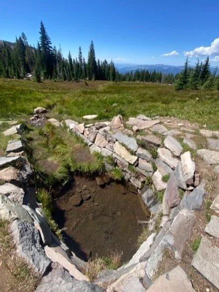 A partially built stone structure in a grassy, mountainous landscape with evergreen trees and blue sky.