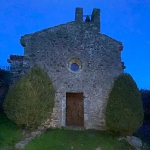 Old stone church with wooden door and two neatly trimmed bushes in front, against a blue evening sky.