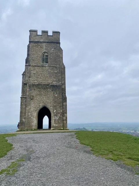 A medieval stone tower on a hilltop with a gravel path leading to its entrance and a cloudy sky in the background.
