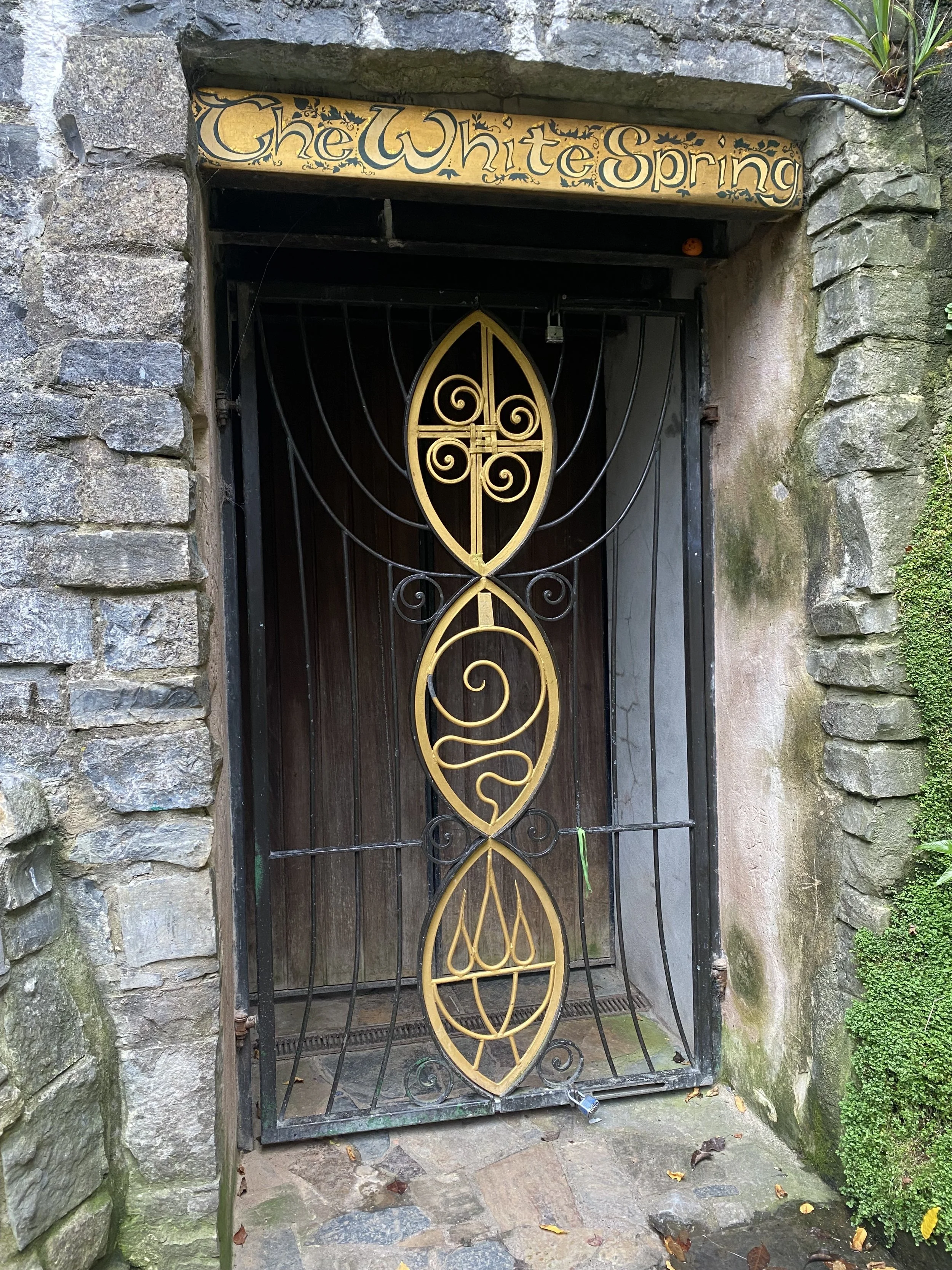 A decorative iron gate with an oval design in the center, secured in a stone archway entrance to a building. Above the gate, a wooden sign reads 'The White Spring' in ornate lettering.