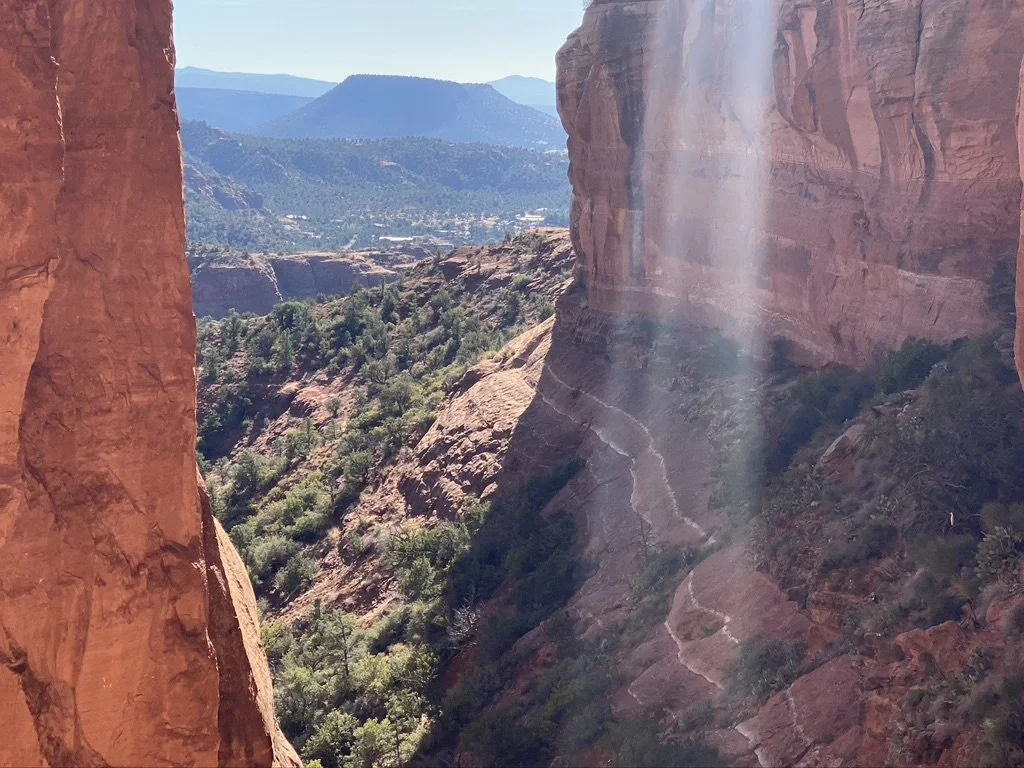 Scenic view of a canyon with red rock formations, sparse green vegetation, and a distant blue mountain range in the background, with sunlight streaming through the rocks.