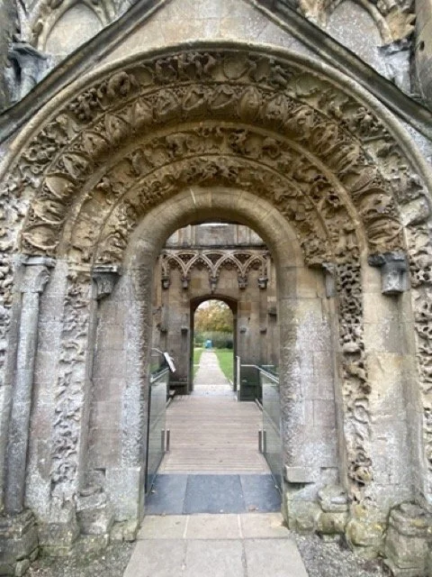 Stone archway with intricate carvings leading to a pathway with a wooden bridge and greenery beyond.