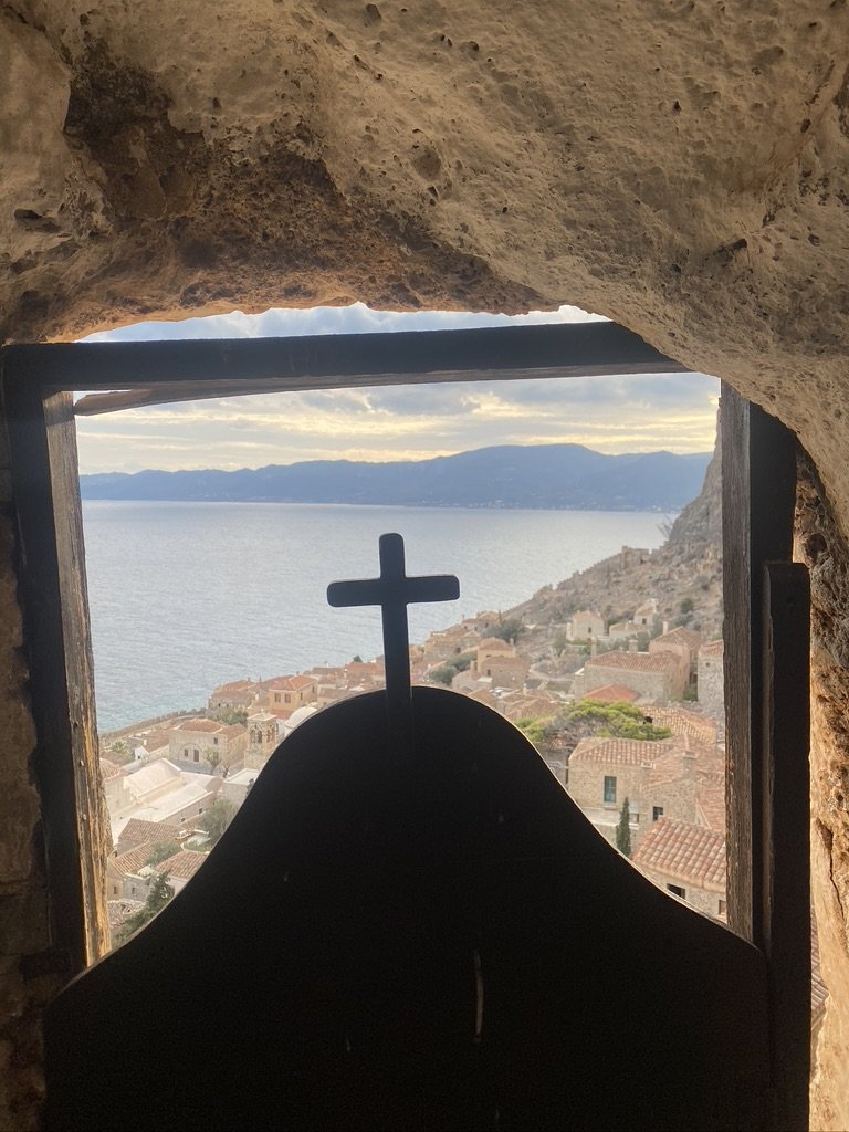 View through a window with a cross, overlooking a coastal village with houses and hills, and the sea in the background.