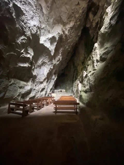 Inside a large cave with rocky walls and wooden benches arranged in rows.
