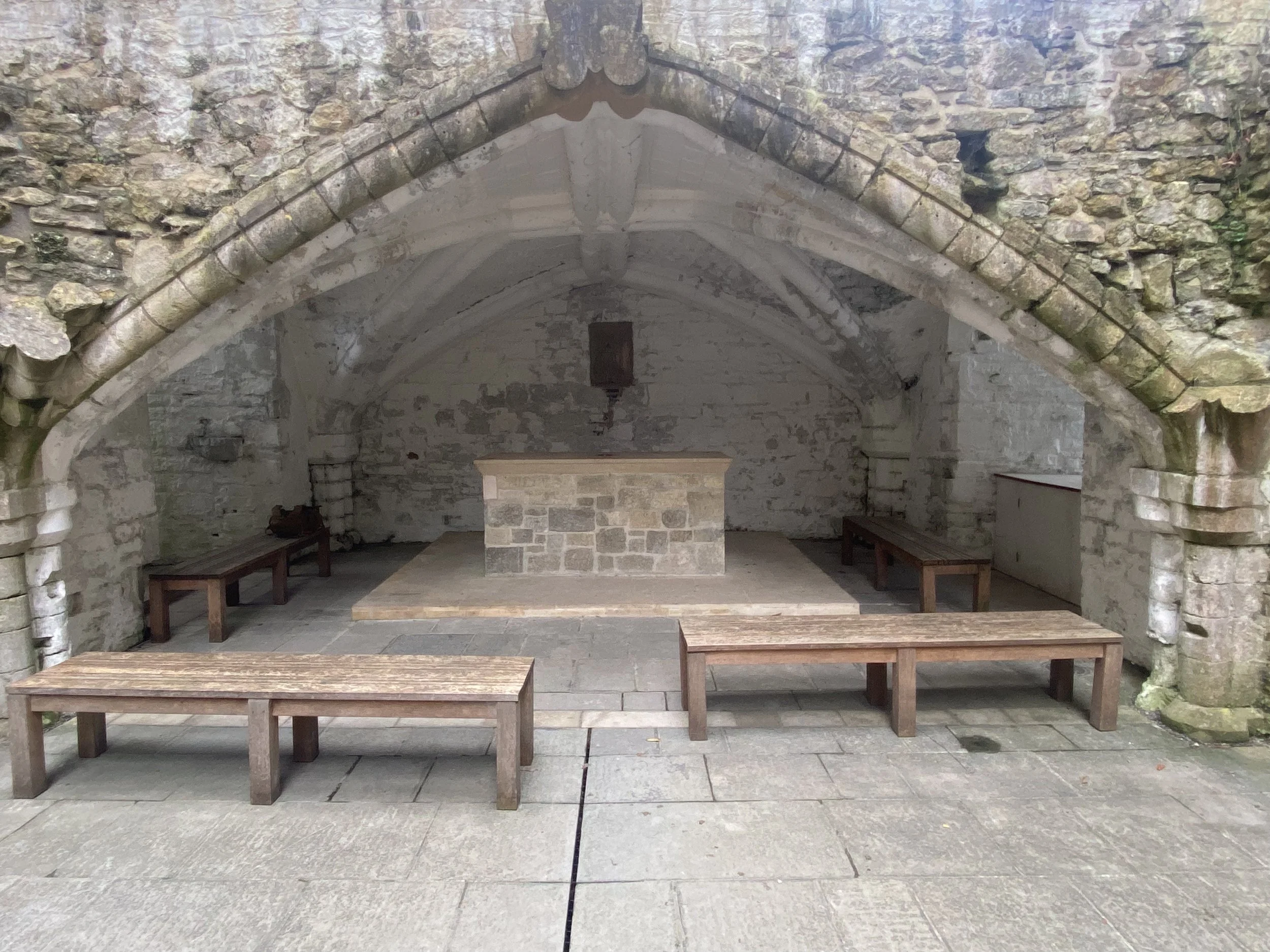 Inside an old stone church or chapel with a central altar, exposed stone walls, and wooden benches for seating.