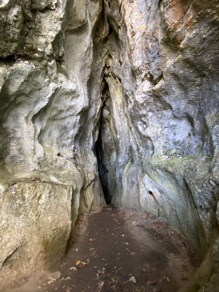 A narrow rocky cave passage with uneven walls and a dirt floor, leading into darkness.