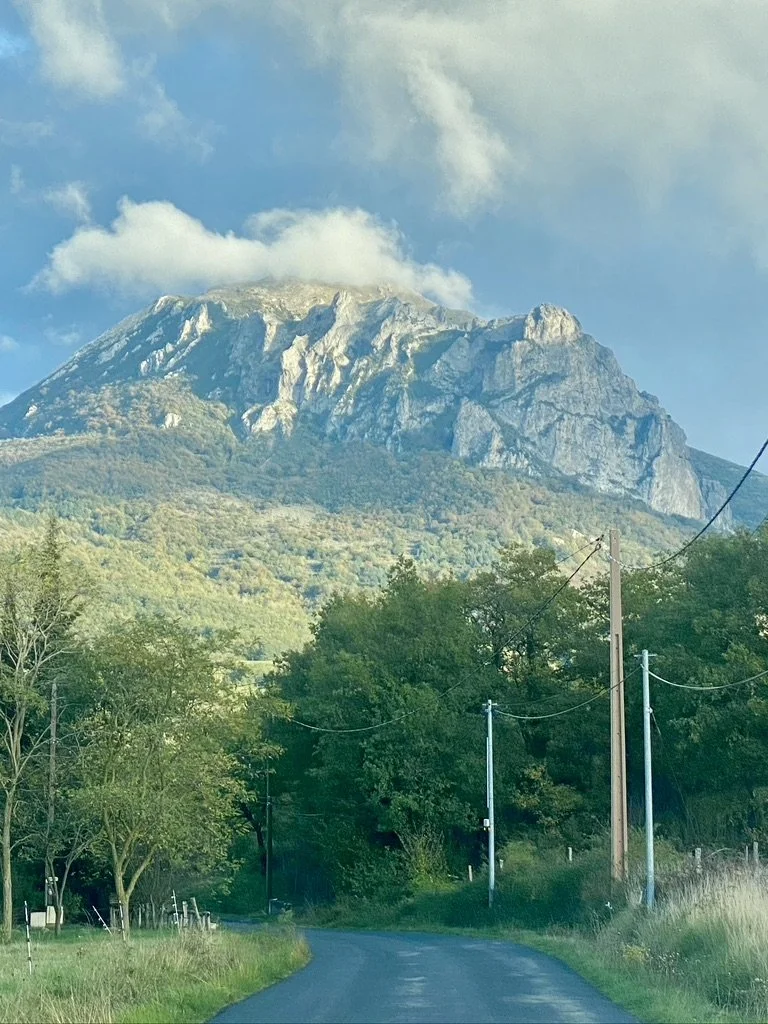 A mountain with rocky cliffs and green forested slopes, with a partly cloudy sky in the background. A winding road with utility poles and wires runs through a lush green landscape in the foreground.