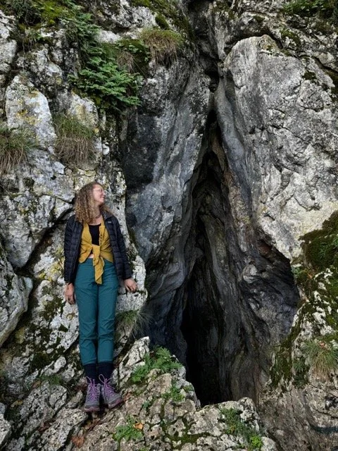 A woman with curly hair standing on rocks next to a large, dark crevice or cave in a rocky, forested area.