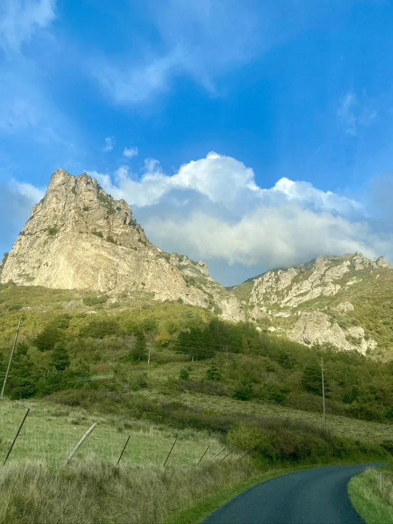 Mountain landscape with rocky peaks, green hills, and a curved road in the foreground under a partly cloudy blue sky.