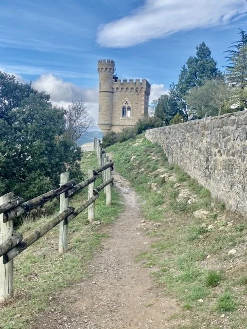 A dirt pathway leading toward a castle-like tower on a hill, with a stone wall on one side and a wooden fence on the other, under a partly cloudy sky.