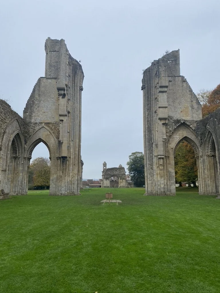 Ruins of an old stone church with large arched windows and tall, broken walls, surrounded by green grass and trees in the background.