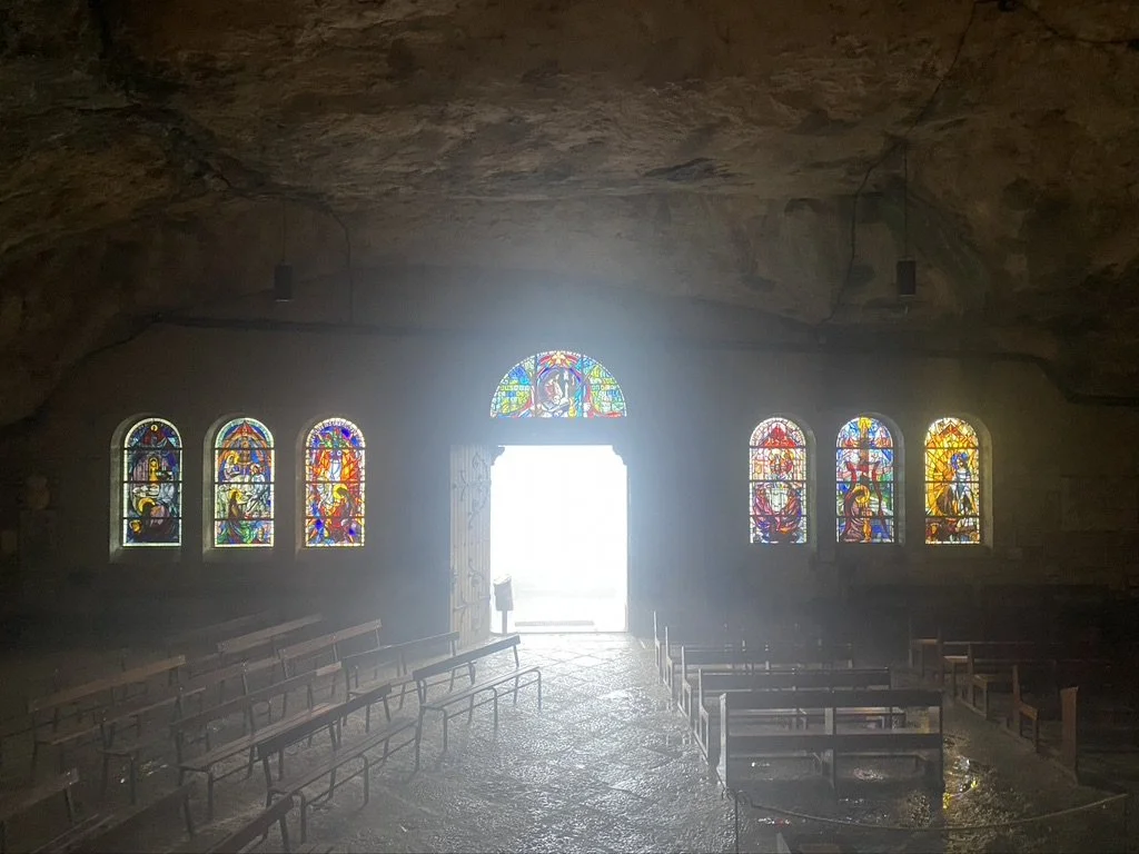Interior of a cave converted into a small church with stained glass windows and wooden pews, sunlight shining through the open door.