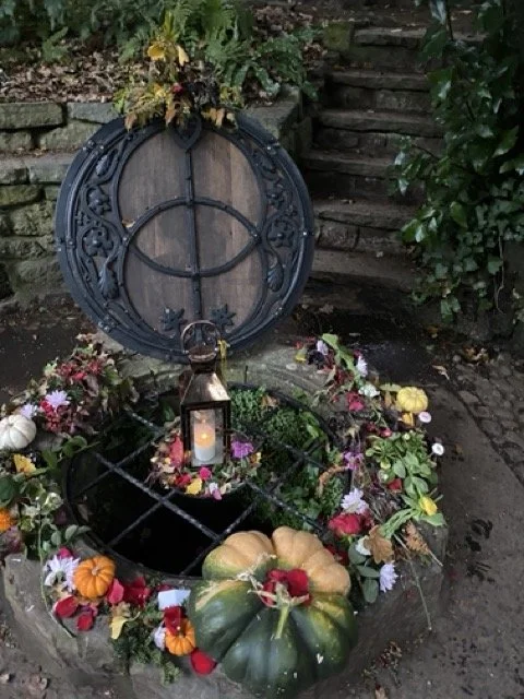 A decorative garden well with an open lid, surrounded by fall flowers and pumpkins, with a lantern inside and stone steps in the background.