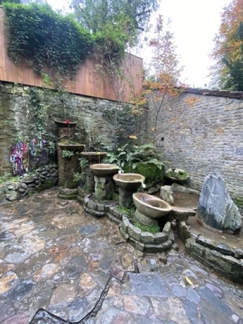 Outdoor stone fountain with multiple basins connected by small waterfalls, surrounded by stone paving and a brick wall in a garden setting.