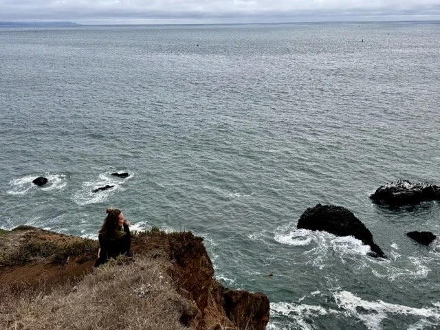 A person sitting on a rocky cliff edge overlooking the ocean with a cloudy sky in the background.