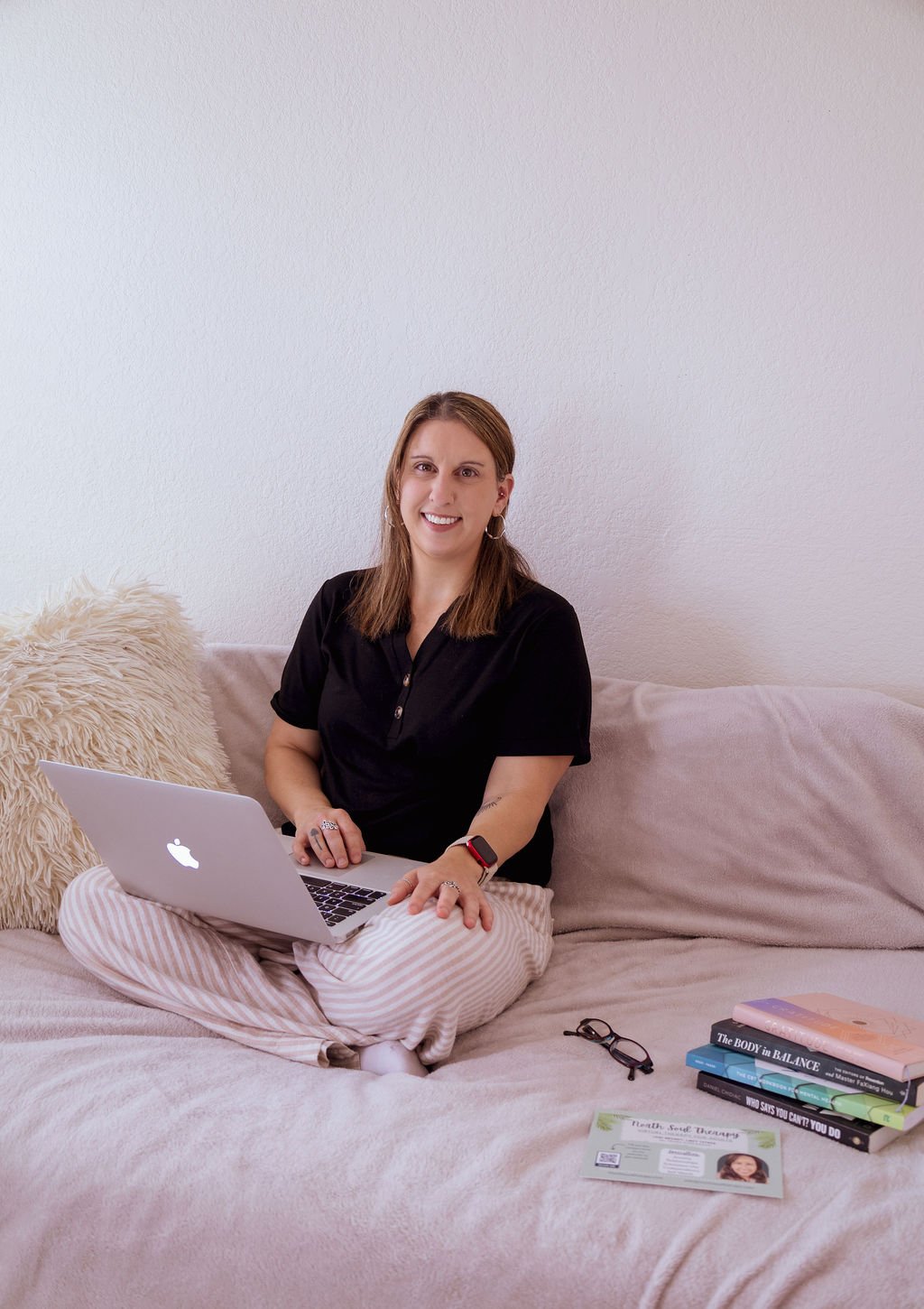 A woman sitting cross-legged on a beige bed with a laptop on her lap. She is smiling and wearing a black shirt and striped pajama pants. Behind her is a fluffy beige pillow, and on the bed are a pair of glasses, a stack of books, and a business card.